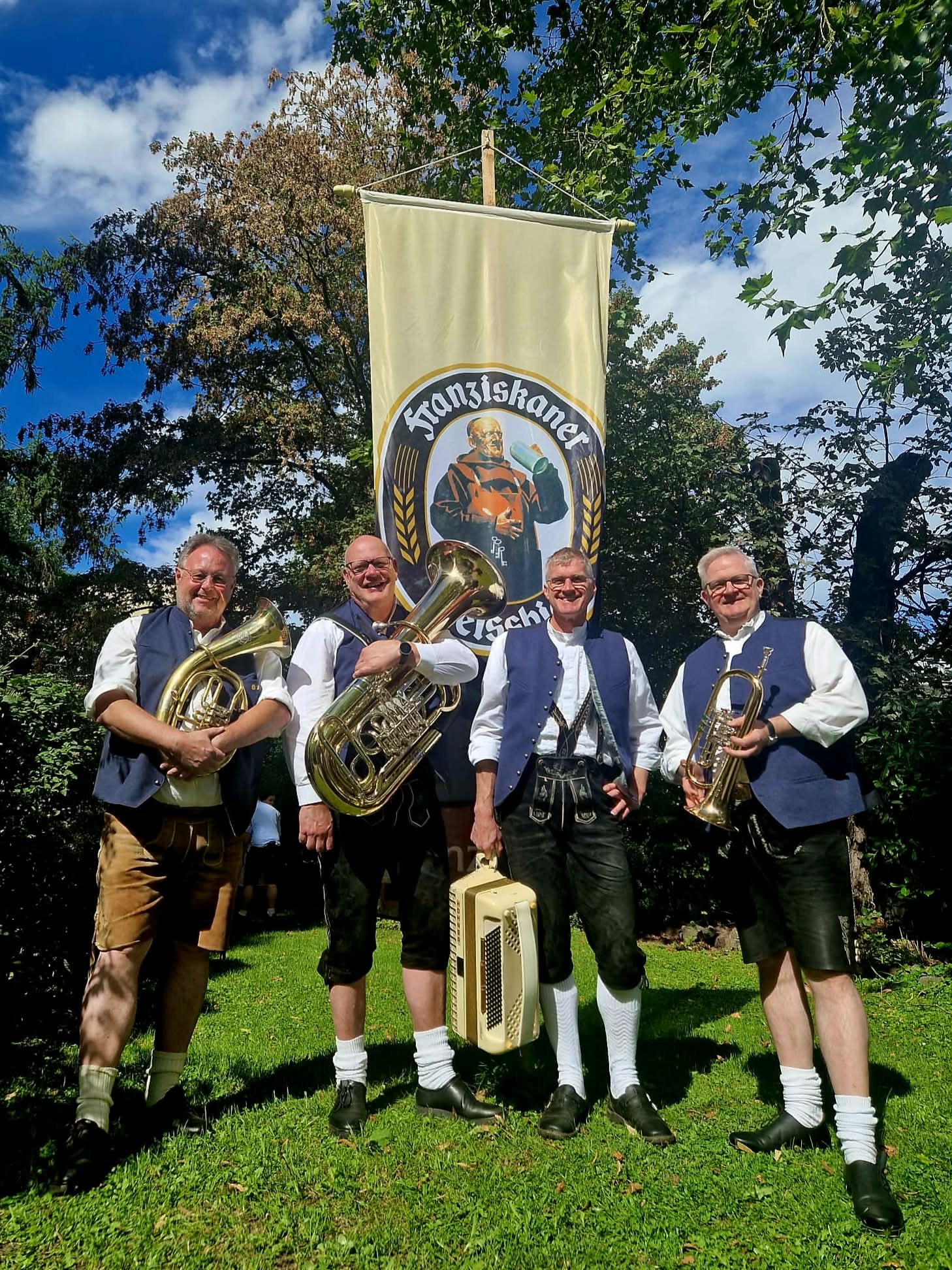 Die vier Wirtshaus-Musikanten in Tracht mit Bassflügelhorn, Tuba, Akkordeon und Trompete auf einer Wiese unter einer Fahne