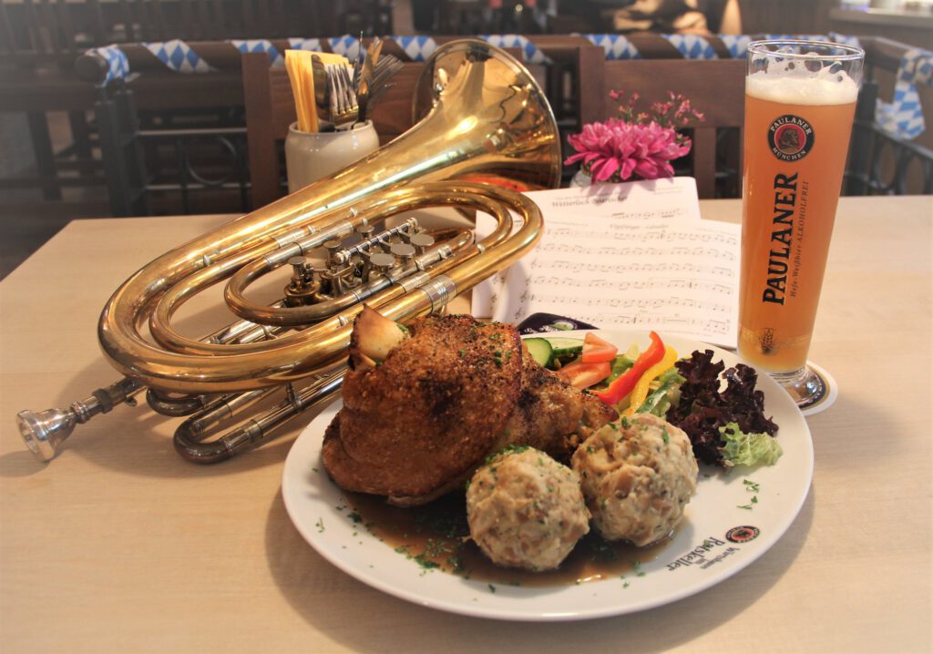 Flügelhorn und Tuba auf einem Wirtshaustisch neben Knödeln und einem Weißbier